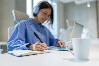 Portatif bilgisayarın başında oturan kulaklıklı gülümseyen şirket çalışanı deftere yazı yazmış.