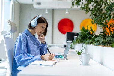 Kulaklıklı ciddi bir ofis çalışanı dizüstü bilgisayara bakarken günlüğe not alıyor.