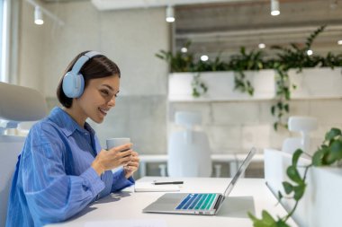 Ofis masasında oturmuş bilgisayarın ekranına bakan kulaklıklı, gülümseyen bir şirket çalışanı.