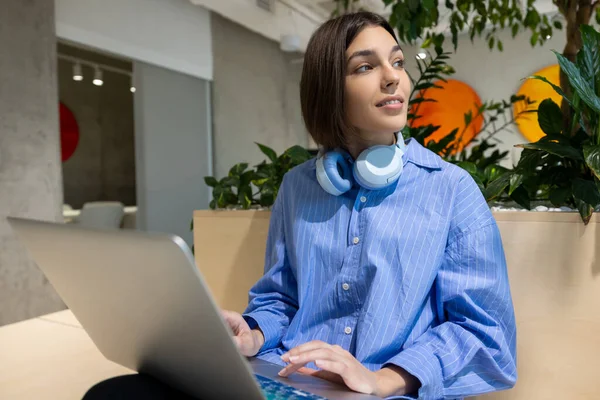 Calm pensive young remote employee tapping on the laptop touchpad while looking into the distance