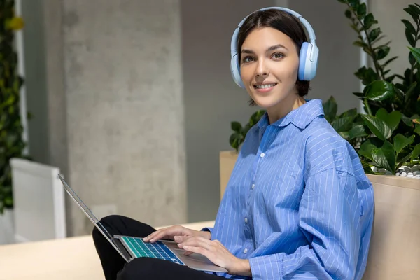ポータブルコンピュータ室内に座っている無線ヘッドフォンで楽しい美しい若い女性を笑顔 — ストック写真