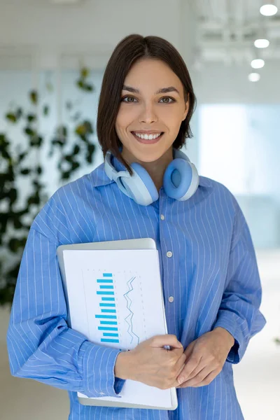 Waist-up portrait of a joyful corporate employee with business documents and the portable computer in her hands standing indoors