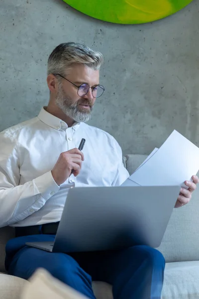 真剣に焦点を当てた男性起業家は彼の手の中に文書を通して見てノートパソコンとソファの上に座って — ストック写真