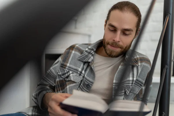 stock image Joyful young man reading book while sitting on stairs in his apartment.