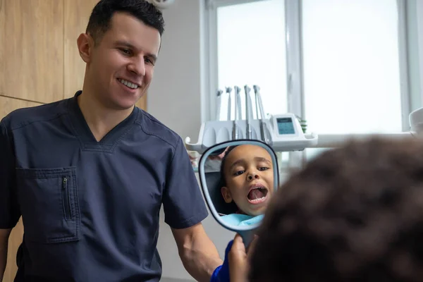 In dentistry. Doctor giving mirror to dark-skinned boy