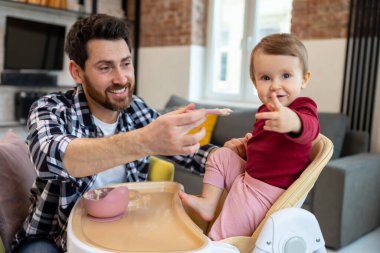 Bakıcı sakallı baba, sevimli bebeğini kaşıkla beslerken modern iç mekanda birlikte oturuyorlar..