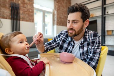 Esmer baba, küçük bebeğine koltukta oturan küçük kızından yulaf lapası yemeyi öğretiyor..
