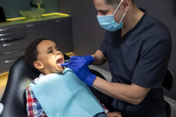 在牙科诊所医生检查他的牙齿时 黑皮肤男孩看起来很害怕 — 图库照片