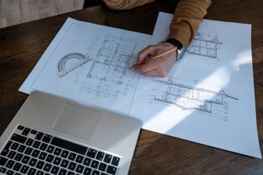 Tanımlanamayan erkek mimar, ofisteki iş yerinde dizüstü bilgisayar ve çizimlerle inşaat projesi çiziyor..