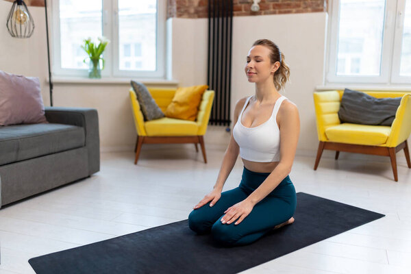 Young fit woman in sportswear practicing yoga on exercise mat in home interior.