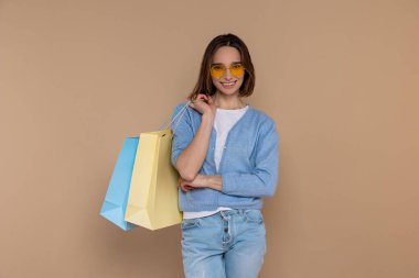 Günlük kıyafetlerle gülümseyen, elinde alışveriş torbalarıyla bej arka planda izole edilmiş kameraya bakan kendine güvenen bir kadın..