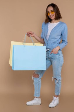 Keyifli, gülen bir kadın, alışveriş torbaları içinde bej arka planda izole bir şekilde moda mağazasında vakit geçiriyor..
