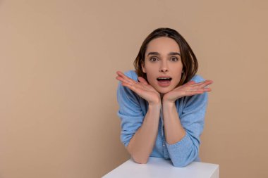 Çene altında elleri olan sıradan giysili bir kadın hayret verici bir şekilde bej arka planda yalnız kaldığını söylüyor..