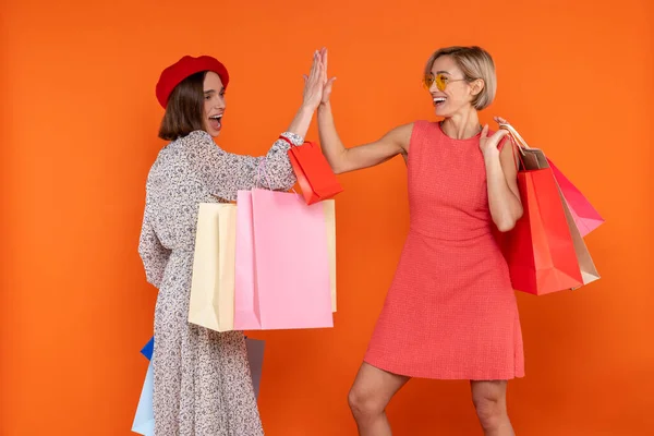 Happy women friends with shopping bags giving high five isolated over orange background.