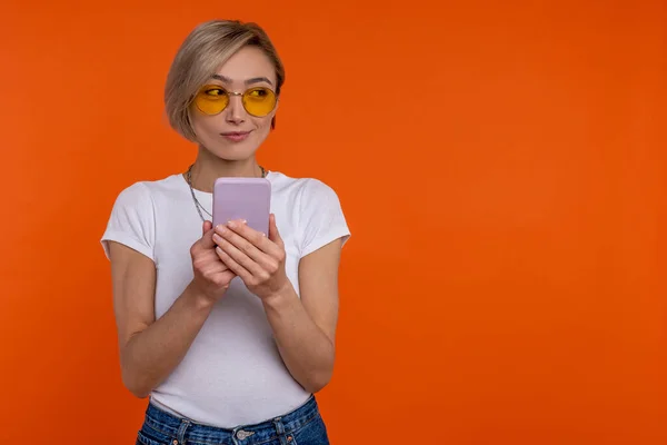 聪明的女人穿着白色T恤 用手机检查社交网络 与橙色背景隔离 — 图库照片