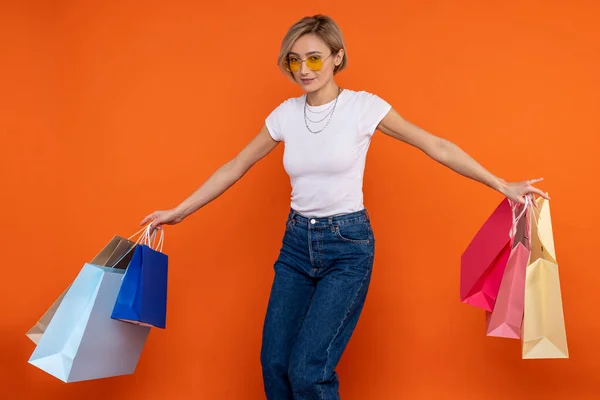 穿着白色T恤和牛仔裤的节日女性提着购物袋 享受独立于橙色背景的购物 — 图库照片