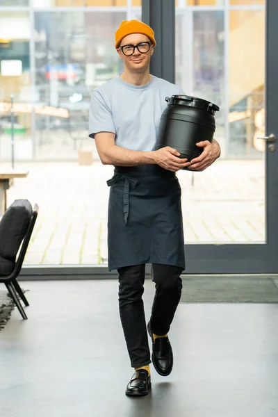エプロンを身に着けている積極的なヒップスターバリスタは コーヒーショップで働くための準備バレルを運ぶ — ストック写真