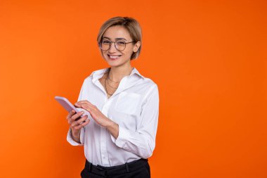 Beyaz resmi tişört giyen kendine güvenen güzel bir kadın turuncu arka planda izole edilmiş akıllı telefon kullanıyor..
