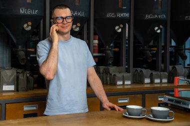 Gözlüklü mutlu barista kahve dükkanında cep telefonuyla konuşuyor, iş yerinde barmen kahve makinesi var.,