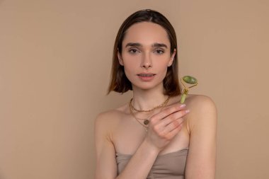 Esmer kadın, bej arka planda tek başına duran cilt bakımı prosedürleri için yüz silindiri kullanıyor..