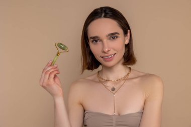 Esmer kadın, bej arka planda tek başına duran cilt bakımı prosedürleri için yüz silindiri kullanıyor..