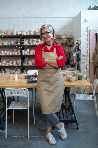 快乐快乐的老年女性成功的自谋职业的陶工穿着围裙坐在桌子后面 手握陶器 对着镜头微笑 — 图库照片