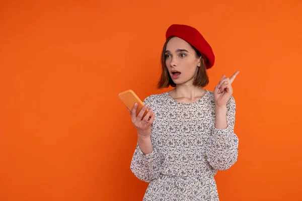 受启发的成年女性穿着雅致的衣服 头戴红色贝雷帽 用手机举起手指 想象着站在橙色的背景下 与外界隔绝 — 图库照片