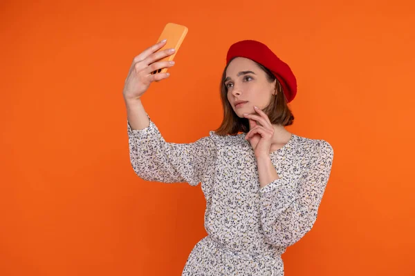 迷人的成年女性 穿着雅致的衣服 头戴红色贝雷帽 手持手机 站在橙色的背景下 显得很有魅力 — 图库照片