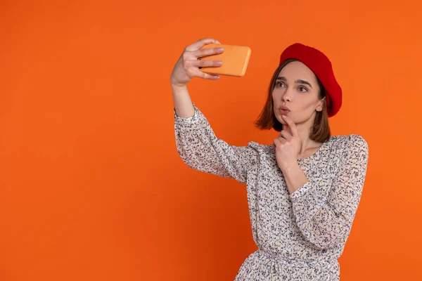 迷人的成年女性穿着雅致的衣服 戴着红色贝雷帽 站在橙色的背景下 在手机上自拍 — 图库照片