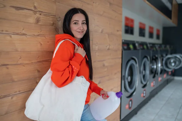 オレンジ色のパーカーを着た若い女性が洗濯機で洗濯洗剤ボトルを持っています — ストック写真