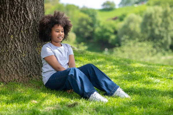 梦中的小女孩穿着白色的T恤衫和牛仔裤坐在树下的草地上 享受着新鲜的头发 — 图库照片