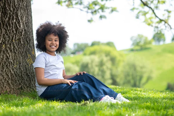 快乐的小女孩穿着白色的T恤衫和牛仔裤坐在树下的草地上 享受着新鲜的头发 — 图库照片