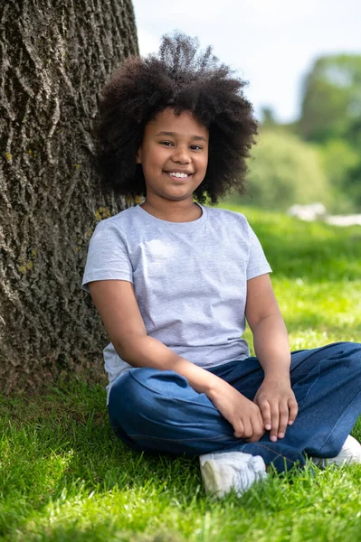 微笑的小女孩坐在树下的草地上 穿着白色的T恤和牛仔裤 在大自然中休息 享受新鲜的头发 — 图库照片