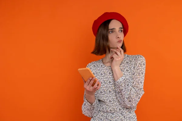 身穿雅致服装 头戴红色贝雷帽 头戴智能手机 站在橙色背景下的多愁善感的成年女性 — 图库照片