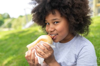 Koyu tenli kadın çocuk parkta çimde otururken zevkle abur cubur yiyor, afro saçlı kız günlük giysiler giyiyor..