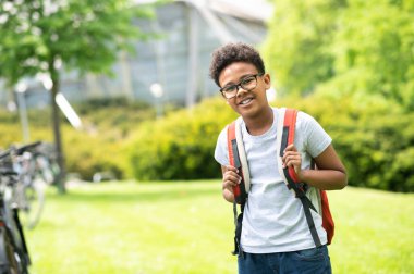 Yeşil parkta duran, sırt çantalı, beyaz tişörtlü, gülümseyen okul çocuğu..
