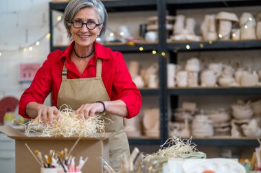 Çömlekçide müşteriler için sipariş edilmiş eşyaları paketleyen yaşlı kadın kil ürünü kutuya koyuyor..
