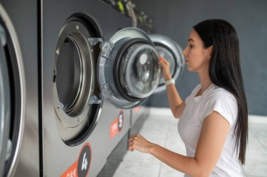 Genç esmer kadın çamaşırhanede çamaşır makinesi kullanıyor.