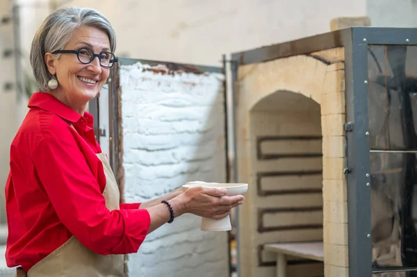 stock image Craftswoman placing clay mugs into kiln for firing pottery while working in workshop, creating earthenware.