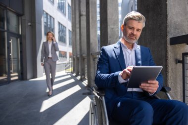 Tekerlekli sandalyedeki başarılı iş adamı elinde tablet bilgisayar, internette geziniyor, dışarıda çalışmak için mobil cihaz kullanıyor..