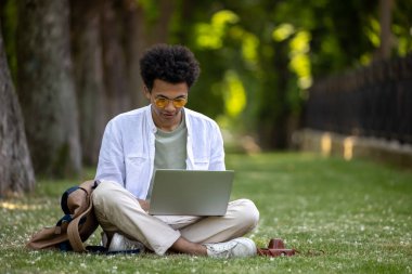Dijital göçebe. Genç kıvırcık saçlı adam parkta dizüstü bilgisayarla çalışıyor.