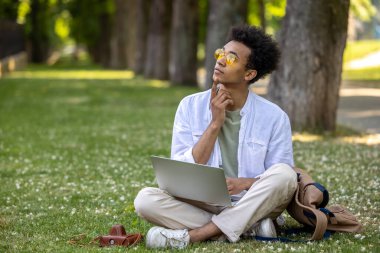 Dijital göçebe. Genç kıvırcık saçlı adam parkta dizüstü bilgisayarla çalışıyor.