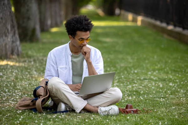 Dijital göçebe. Genç kıvırcık saçlı adam parkta dizüstü bilgisayarla çalışıyor.