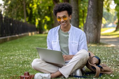 Uzaktan çalışıyor. Hafif giysili bir adam çimenlerde oturuyor ve dizüstü bilgisayarla çalışıyor.