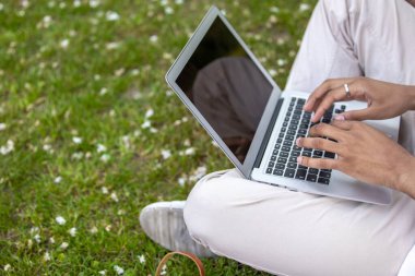 Uzaktan çalışıyor. Hafif giysili bir adam çimenlerde oturuyor ve dizüstü bilgisayarla çalışıyor.