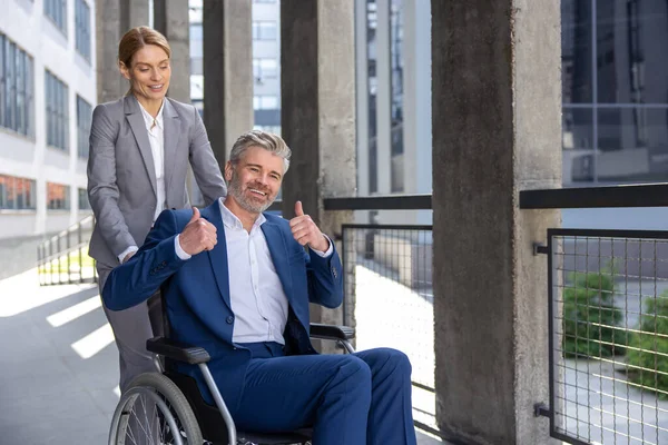 Blonde woman assisting man business partner in wheelchair going to office, male showing thumb up, like gesture.