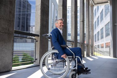 Gülümseyen gri saçlı engelli adam tekerlekli sandalye şehrinin arka planında resmi bir takım elbise giyiyor..