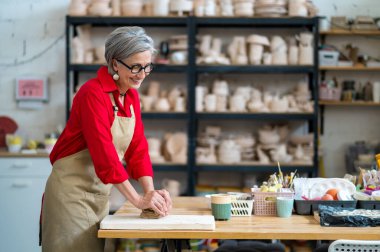 Çömlekçilikte elle yoğurma tekniği. Olgun bir kadın, stüdyoda masanın arkasında elleri killi bir şekilde bekliyor..