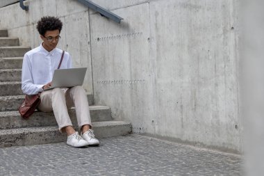 Uzaktan çalışıyor. Beyaz tişörtlü kıvırcık saçlı genç adam merdivenlerde dizüstü bilgisayarla oturuyor.