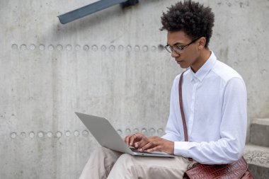Uzaktan çalışıyor. Beyaz tişörtlü kıvırcık saçlı genç adam merdivenlerde dizüstü bilgisayarla oturuyor.